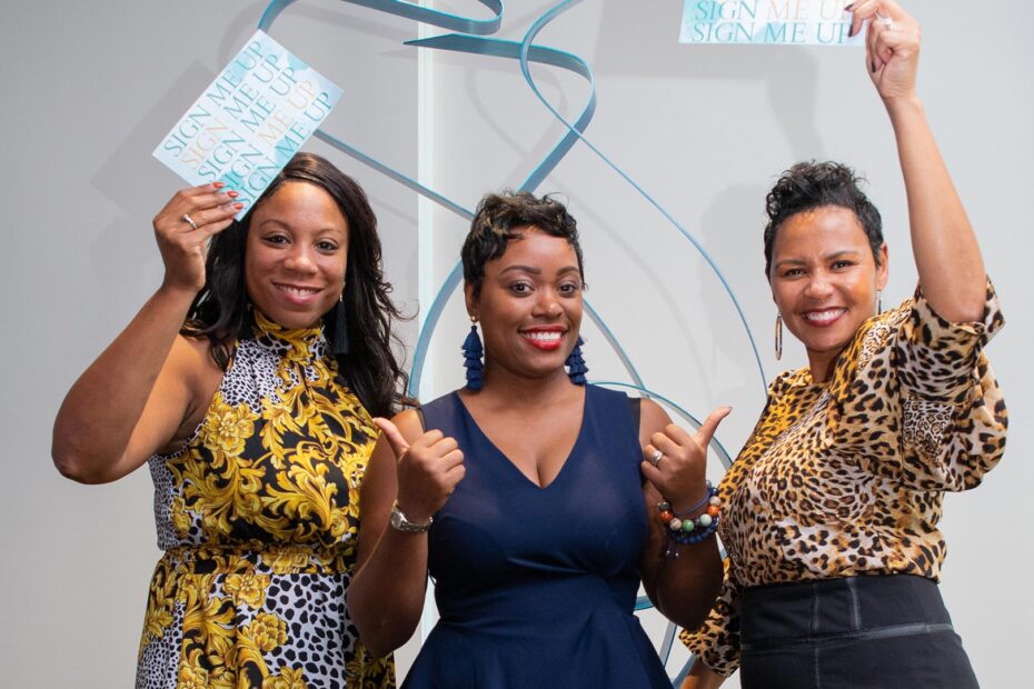 Three smiling women holding "sign me up" cards at a Women's Committee event