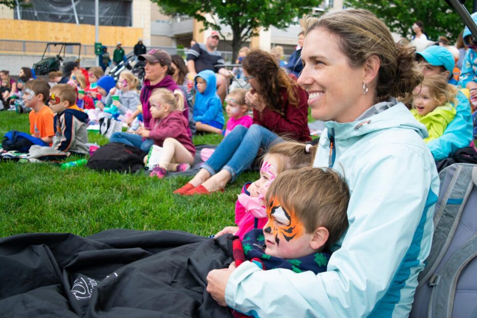 A mother with two children wearing face paint sit it an audience on a grassy lawn.