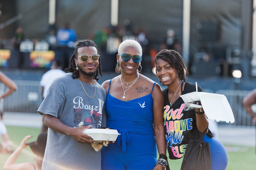 Three people smiling and looking at the camera. They are dressed casually. They are standing in the audience of a crowd gathered to listen to music on a stage in the distance.