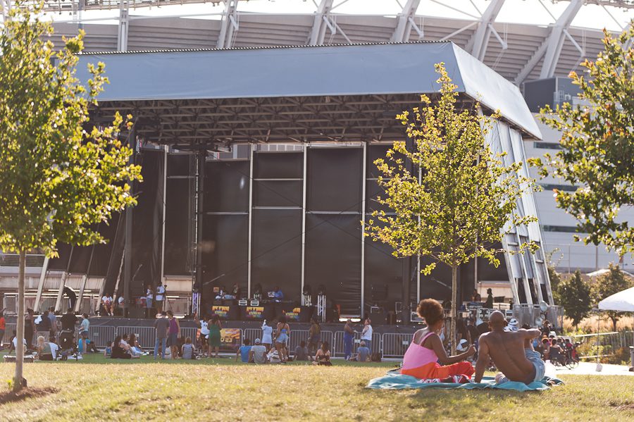 A group of people facing an outdoor stage. There are people on the stage with equipment so they can play music.
