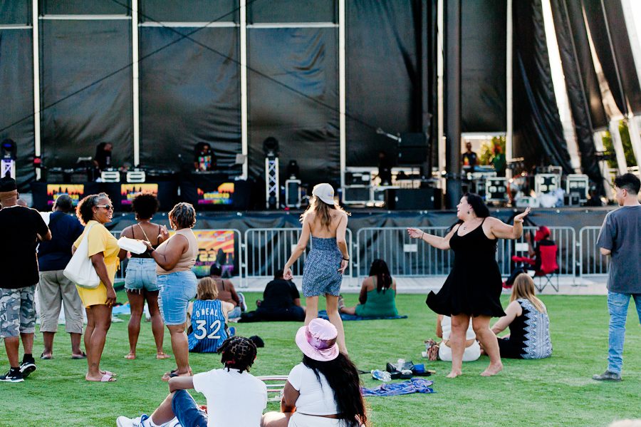 A crowd of people in front of a outdoor stage. They are listening to music from DJs and dancing.