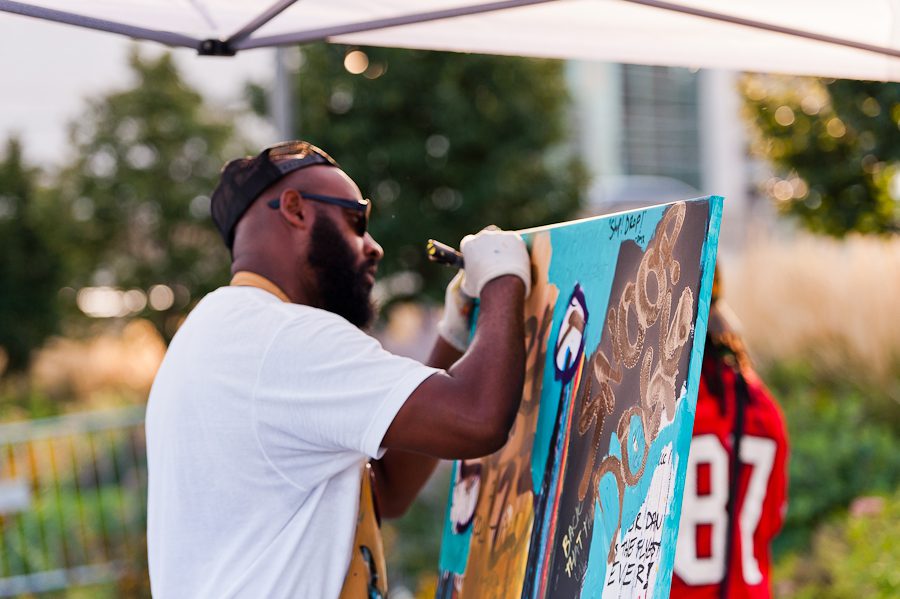 An artist working on a large painting. He is working outside. There are trees and plants behind him.